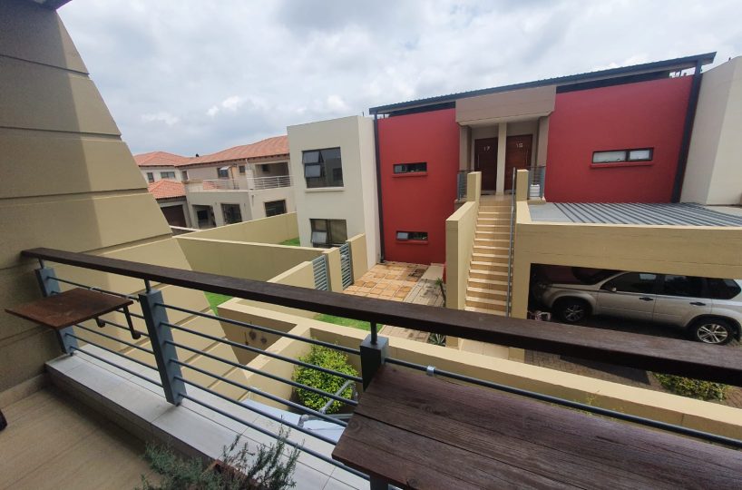 View from the bacony of a townhouse that is for sale in The Villas, Glen Eagle Estate, Kempton Park. It looks out onto the neighboring block of 4 townhouses, with stais and the carport. The block is painted red and 3 neutral colors. The small kitchen windows are visible.