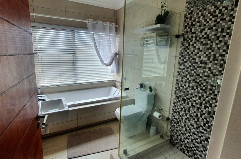 Full bathroom with modern finishes. A big rectangular bathtub under a big window on the far wall. A big, glass shower with mosaic tiles on the wall. Interior of a townhouse that is for sale in The Villas, Glen Eagle Estate, Kempton Park.
