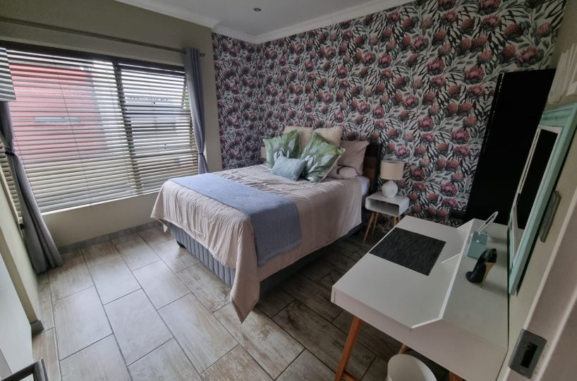 Main bedroom of a townhouse that is for sale in The Villas, Glen Eagle Estate, Kempton Park. It has an accent wall with protea wall paper that is pink, green and white. Tiled floor, a double bed, a bedside table and a dressing table with a mirror. A large window with blinds and curtains.